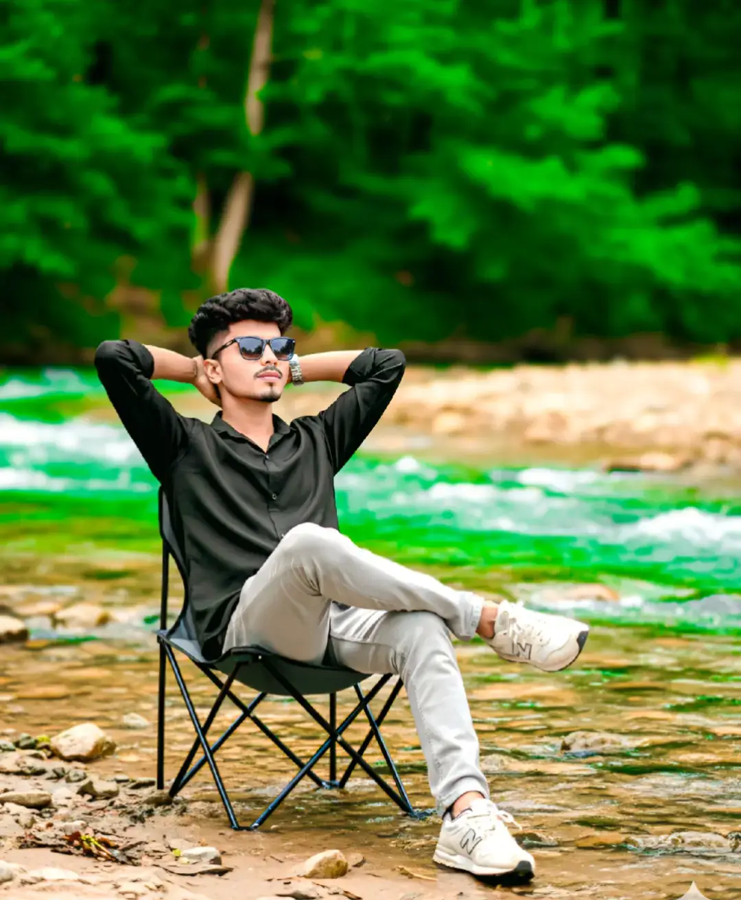 Ultra-realistic photo of a man (same face and hairstyle as reference) wearing sunglasses, relaxing in a camping chair by a running riverbank. He has his hands behind his head and legs crossed, wearing a full sleeve black linen.shirt, grey trousers, half white new balance sneakers. He wears a silver chain watch. /-#Outfit cowok berwarna #fyp