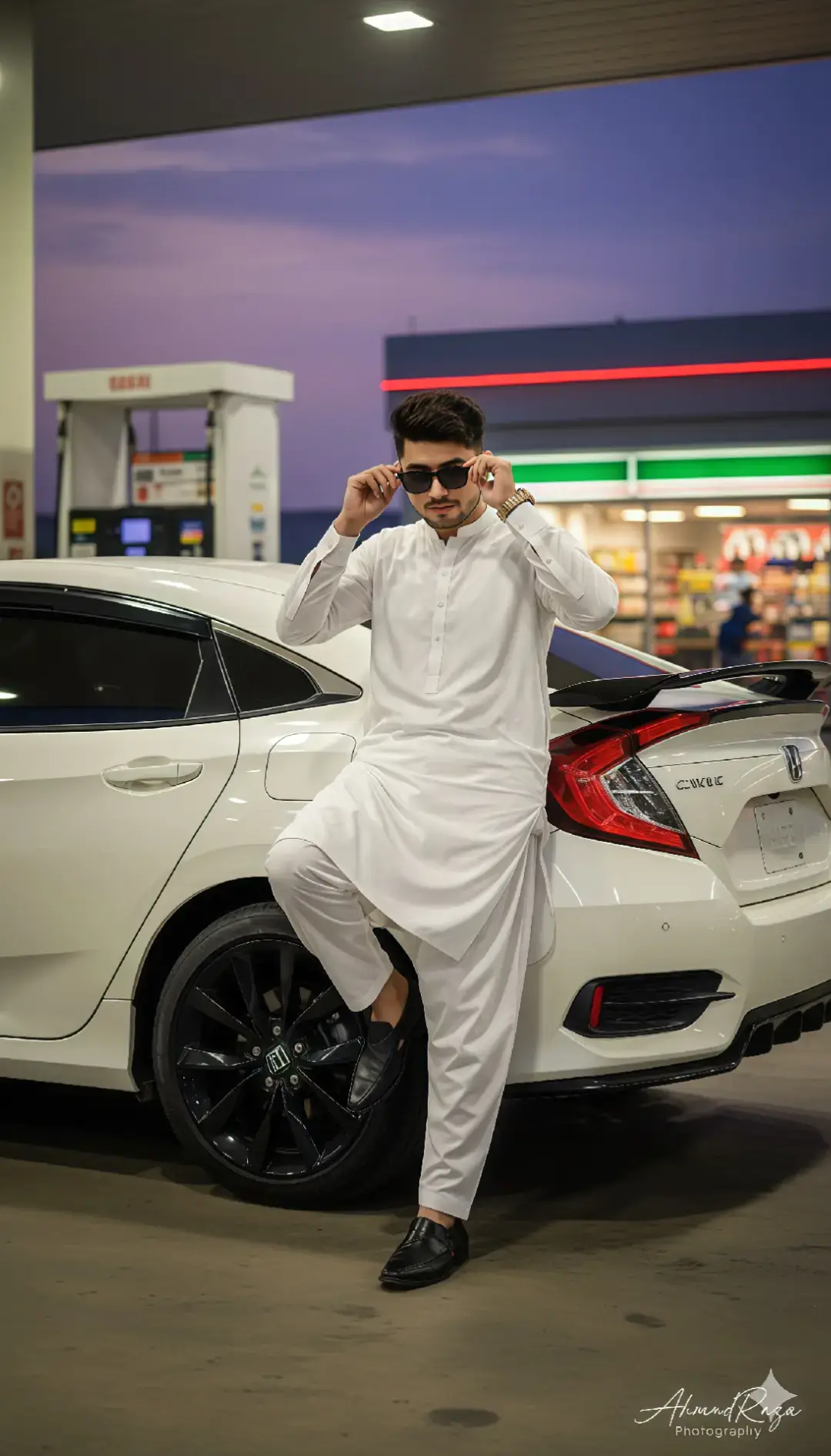A stylish young man standing beside a white Honda Civic at a gas station during evening twilight. The man is wearing a traditional white shalwar kameez with black formal shoes and black sunglasses. He is posing confidently, one hand touching his hair. The Honda Civic has a rear spoiler, black alloy rims, and tinted windows. The gas station has bright lights, a fuel pump beside the car, and a convenience store glowing softly in the background. The sky shows a purple-blue sunset gradient. The overall look is cinematic, clean, and modern — professional portrait photography style, shallow depth of field, soft background blur, realistic lighting, HDR tone, crisp details, fashion photoshoot aesthetic. —v 5 —ar 2:3 —style photo —lighting: evening, cinematic, natural. 