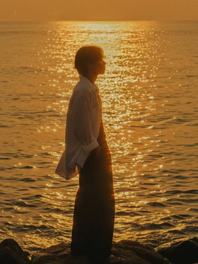 Prompt : Based on the referenced face and body, create a wide cinematic surreal shot in either vertical or horizontal format: the atmosphere is nostalgic, melancholic, yet peaceful. The character is posed in a relaxed 3/4 angle, expressing a sense of enjoyment under the sunset by the sea. Strong highlights (reflections shimmering on the water surface) dominate the lighting. The primary color palette is warm golden tones, almost monochromatic. Film grain texture and halation (a soft bloom around the light) give the image a vintage, dreamlike quality. The character wears a loose white shirt with its tail flowing in the wind and wide-legged black trousers, standing on rocks near the sea (the character occupies only about 20% of the overall scene, appearing small within the vast environment). Full-body framing, standing tall, balanced on one foot/rock — posture calm and relaxed. The face is obscured by backlighting in one interpretation, yet in another version the facial details remain visible. The surrounding scenery is a vast, boundless sea — empty except for water, the subject, and the rocks. The moment captured is at sunset or sunrise, with light spreading near the horizon. Framing: vertical or horizontal composition, with the subject placed near the center of the frame or slightly above — horizon positioned toward the upper part of the frame. The camera is at eye level or slightly lower than the subject, evoking the feel of a medium–telephoto lens (compressed perspective), which causes the background to collapse and the shimmering reflections on the water to stretch into elongated streaks. Visual leading lines: the sparkling highlights on the water guide the viewer’s gaze toward the subject. pria tersebut duduk dengan tangan yang bersandar pada lutut nya menghadap sunset seolah membelakangi kamera. #gemini #ai #geminiedit #google #geminiaitutorial 