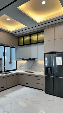 Kitchenset berbentuk L-shape memadukan warna abu-abu doff dengan top table marble putih urat abu, menciptakan tampilan bersih dan elegan. Kabinet atas memakai kombinasi pintu kaca hitam dengan lampu LED warm yang memperkuat kesan premium. Bagian island table menggunakan marble rounded corner dengan aksen list kayu vertikal, berfungsi ganda sebagai area makan santai dan pemisah ruang. Di atasnya tergantung tiga lampu gantung silinder hitam yang memberi pencahayaan dramatis namun tetap hangat. Lantai menggunakan homogeneous tile marble glossy, memantulkan cahaya dari plafon drop ceiling berlampu warm white, sehingga ruangan terasa luas dan berkelas. Perpaduan warna grey, putih marble, dan hitam doff membuat dapur ini terlihat modern, rapi, dan mewah — a perfect balance between function and style. #CapCut 