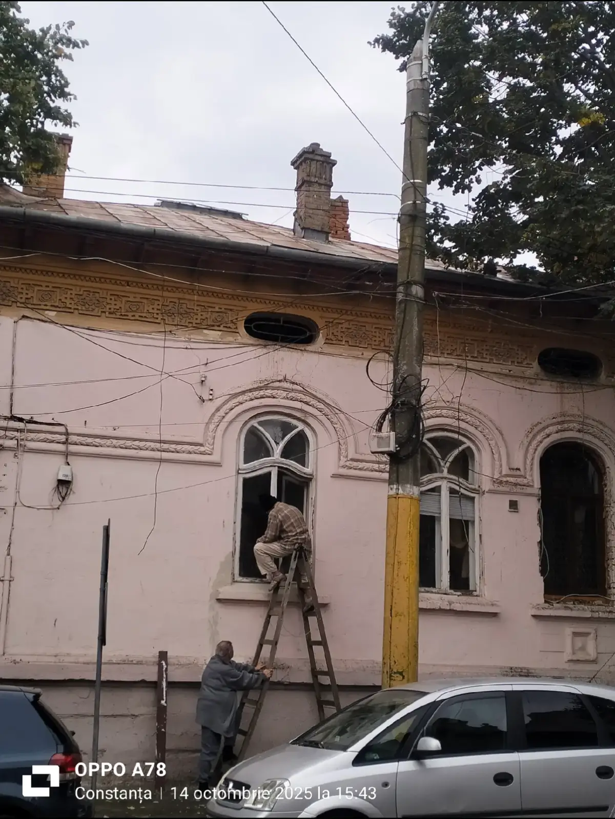 Timpul trece peste noi, dar și peste lucruri. Își lasă amprenta peste tot. Diferența este că noi, oamenii, putem avea grijă de ele — le putem proteja, le putem prelungi viața, chiar mai mult decât pe a noastră. Ele, în schimb, nu pot avea grijă de noi. Unii oameni au însă un rol aparte: acela de a conserva și de a prelungi viața obiectelor , clădiri, artă, natură, unelte , dincolo de propria lor durată de viață. Prin grijă, restaurare și mentenanță, ei transmit generațiilor viitoare moștenirea materială a lumii în care trăim. Strada 1907 e plină de clădiri vechi. Unele nu mai sunt decât niște urme, altele par gata să cadă la primul cutremur, iar câteva încă sunt locuite. Ieri, trecând pe acolo, am avut o surpriză: la o casă pe care o mai admirasem de multe ori, am văzut doi prieteni trecuți de 60 de ani cum pregăteau geamurile pentru iarnă. Unul ținea scara, iar celălalt, cocoțat cu un șpaclu, răzuia vopseaua scorojită care se desprindea la o simplă atingere. Când i-am văzut, i-am fotografiat instinctiv. Mi-am dat seama imediat ce fac și m-am bucurat. M-am apropiat, i-am salutat și i-am felicitat. Se uitaseră la mine și chicoteau, amuzați că îi pozam. Pe fața mea se citea bucuria , bucuria de a vedea doi oameni, poate doi bunici, care alegeau să-și salveze bucățica de casă cu propriile forțe, fără să o lase în paragină. Știu că este o casă veche și că nu au voie să pună termopan, ci doar lemn. Respectul lor pentru reguli, pentru autentic, și felul în care vorbeau despre casă mi-au stârnit o emoție puternică. M-am simțit mândră că i-am cunoscut. Casa e mare. Vecinul de lângă și-a pus termopan. Ei nu vor. Poate, dacă lemnul va ceda, îl vor înlocui tot cu lemn. De la Primărie nu au primit ajutor de peste 40 de ani, în afară de un strat grăbit de var aruncat peste fațadă. Casa poartă cicatricile vremii,urme care trebuie refăcute de un specialist, dar care vorbesc despre timpul trecut. La insistențele domnului, am intrat în casă. Nu m-a lăsat să mă descalț și m-a primit ca pe o cunoștință veche. Ușile camerelor urmează să fie vopsite, iar iluminatorul din living pare din altă lume , întreținut tot de el. O casă veche, norocoasă, cu inima caldă, încă în picioare și plină de viață. Le-am mulțumit din suflet și am plecat fericită. Le-am spus că i-am fotografiat din spate , au zâmbit și mi-au spus că nu-i nicio problemă. I-am lăsat exact cum i-am găsit: lucrând. Două ore mai târziu, am trecut iar pe acolo. Erau tot acolo, la altă fereastră. Lăsarea serii nu părea să-i deranjeze. Un refuz al soluției rapide, în favoarea celei corecte din punct de vedere istoric. Au ales să păstreze trecutul cu resurse proprii și cu mult suflet, chiar dacă înseamnă muncă grea, făcută de mâini care au trudit deja o viață. O fac în fiecare an. Și, în felul lor, țin timpul pe loc. #moștenireculturală #restaurarecuinimă #clădirivechi #grijășirespect #româniideaur 