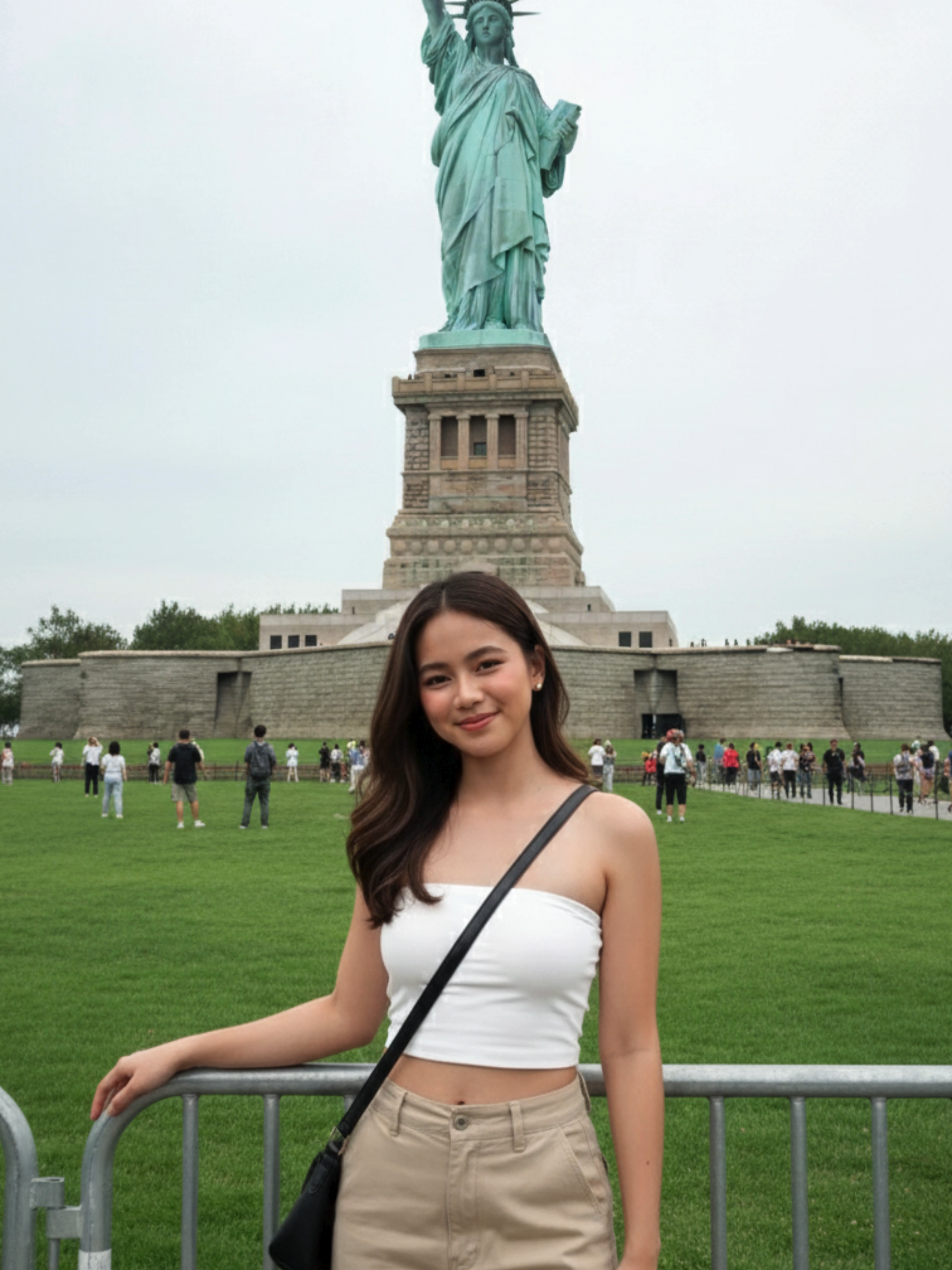 Gemini AI prompt: Three-quarter length portrait of me (preserve my facial features i should be recognizable) standing on Liberty Island lawn in front of the Statue of Liberty, shot on iPhone 15 Pro in Portrait mode with Natural Light effect, bright overcast daylight creating soft even illumination, iconic Statue of Liberty copper statue with oxidized green patina towering in background on tan granite and concrete pedestal atop Fort Wood's star-shaped base with eleven-point star fortification walls visible, woman wearing white strapless crop top and tan beige cargo pants, black crossbody bag, relaxed standing pose with hand resting on silver metal crowd control railing, soft natural smile, bright green manicured lawn surrounding pedestal plaza, other tourists visible in background on upper observation areas, grey-blue granite walls of historic Fort Wood star fort visible at base, overcast sky creating diffused natural lighting without harsh shadows, casual tourist moment aesthetic, metal safety railings in foreground, shot with iPhone computational photography with natural background depth separation keeping statue sharp while subject is primary focus, bright airy color grading typical of overcast daylight conditions, photorealistic quality with modern smartphone characteristics. Do not over-edit or enhance the face in any way. Maintain a high degree of facial fidelity to the provided image.