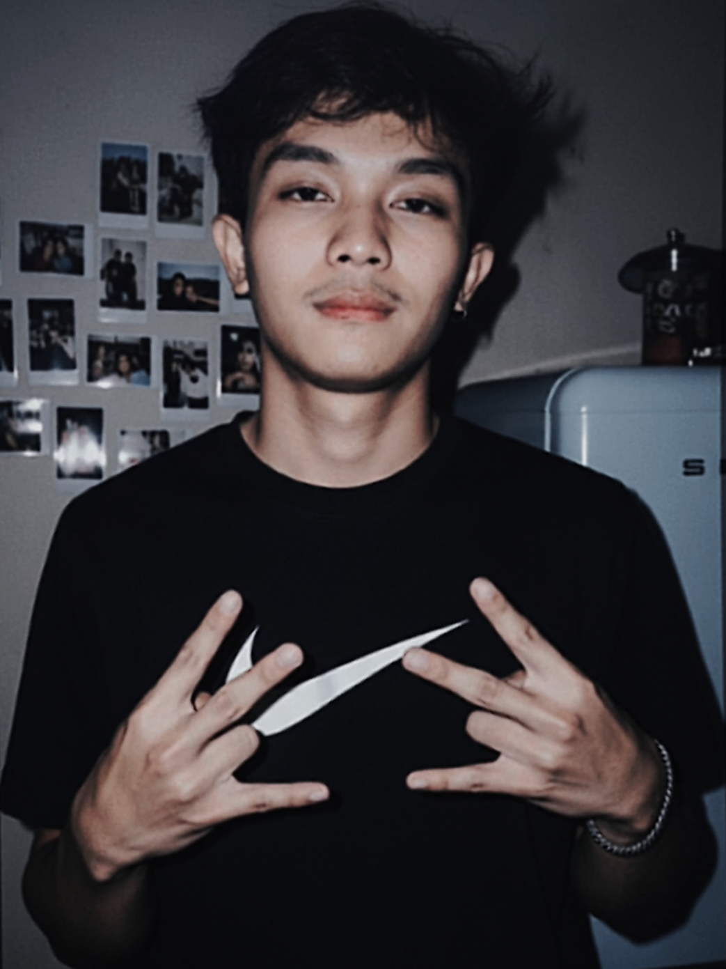 Ubah foto yang saya unggah tanpa mengubah wajah dan rambut, seorang pemuda berdiri tegak dengan rambut sedikit berantakan terkena angin, menghadap kamera, mengenakan kaus Nike hitam dengan logo putih, membuat gerakan tangan hip-hop. Latar belakang dalam ruangan dengan foto polaroid dan kulkas biru. Ditembak dengan flash, estetika retro Y2K 90-an, komposisi yang sama dengan gambar referensi. #promt #gemini #ai 