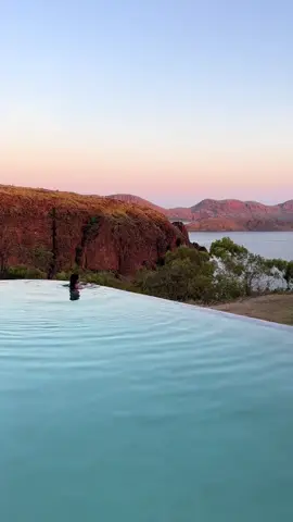 Dive into the beauty of the Kimberley 🏊‍♀️ When it comes to cooling off with a view, it doesn't get much better than wading into @Discovery Parks & Resorts infinity pool overlooking Lake Argyle. 🎥: @Abs  📍: Discovery Parks & Reaorts Lake Argyle, @Australia's North West, @Western Australia  #SeeAustralia #ComeAndSayGday #TravelTok #Travel #Holiday ID: A person swimming in an infinity pool overlooking red rocky peaks and a calm blue lake,