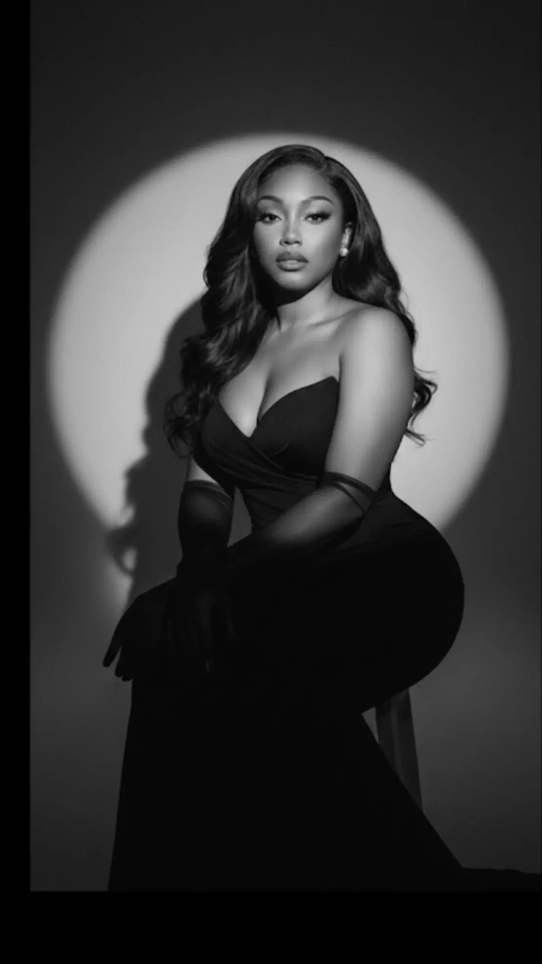 Prompt : Without changing any of my facial features and Appearances Using my picture in a glamorous woman in a strapless black evening gown, sitting gracefully against a dramatic circular spotlight backdrop. She wears sheer black gloves and minimal jewelry, exuding elegance and poise. The image is in black and white, with cinematic lighting and deep shadows, creating a timeless, vintage-inspired fashion editorial look.
