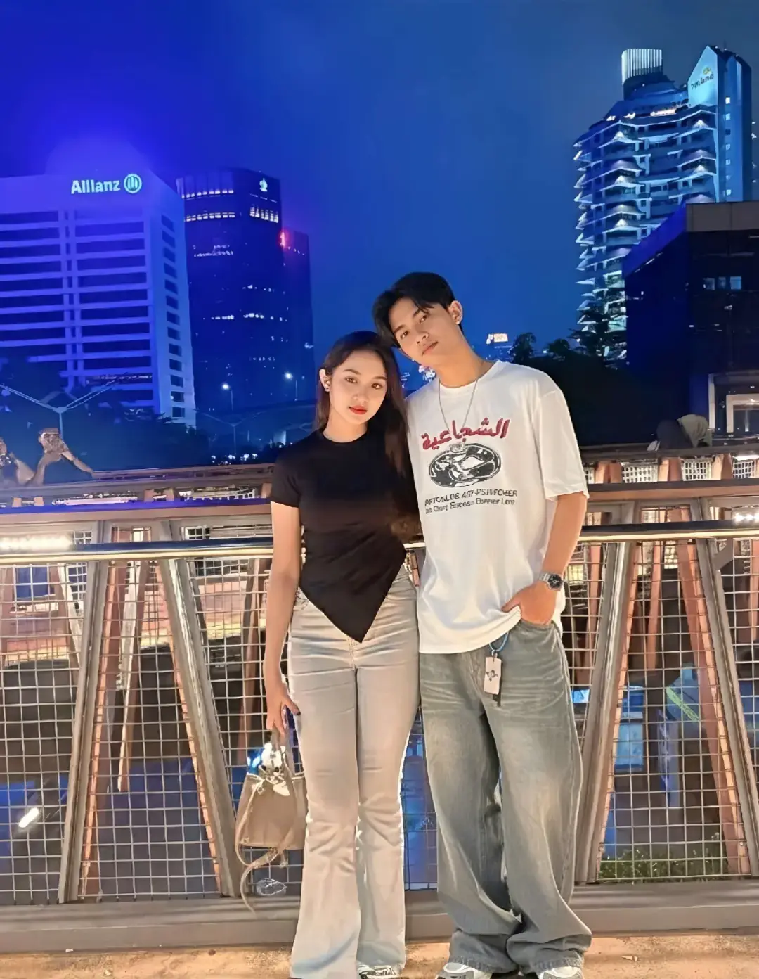 TUTORIAL?? CAPTION‼️ 1. Buka google/apk gemini di hp kalian 2. Masukan photo wajah kalian dan pastekan  prompt:👇🏻 Coba buatkan sebuah foto potret definisi tinggi yang menawan dari sepasang kekasih muda romantis yang sedang berpose di sebuah jembatan penyeberangan modern di tengah pusat kota yang ramai saat malam hari dari dua subjek di atas. Wanita muda, dengan rambut hitam panjang bergelombang yang jatuh indah, mengenakan atasan hitam lengan pendek dengan potongan asimetris yang unik dan flare jeans berwarna abu-abu muda yang trendi, sambil memegang tas tangan kecil berwarna krem. Pria di sampingnya, dengan gaya kasual namun stylish, mengenakan kaus putih oversize dengan cetakan grafis berhuruf Arab yang khas, celana baggy jeans biru pudar, dan chunky sneakers modern. Ia menyandarkan kepalanya dengan lembut ke kepala wanita, menampilkan momen keintiman dan kasih sayang. Latar belakang foto ini adalah pemandangan kota di malam hari yang spektakuler, didominasi oleh gedung-gedung pencakar langit yang menjulang tinggi, termasuk gedung 