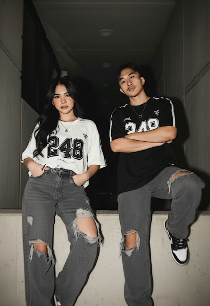 Prompt Gemini ai gaya vintage terbaru. Buat foto ini tanpa merubah wajah, hidung, bibir 100% harus mirip asli menjadi foto ,Gaya fotoristik vintage resolusi 4k. Pria dan Wanita berpose dengan percaya diri dalam posisi duduk  yang sedikit kental, sedikit menjulurkan kaki kanannya kedepan. dan 1 tangan wanita seperti sedang memegang hp berwarna putih atau cream dengan bulatan di casenya dan memiliki 2 kamera. Wanita dan pria mengenakan pakaian yang ada di foto ke 2 dan mengenakan sepatu Air Jordan . Dia mengikat kacamata hitam Prada ke rambutnya dan rambut hitam sangat panjang butterfly sedikit gelombang ujungnya. biarkan dia memakai di atas bahu dan bingkai wajahnya . perhiasan itu beberapa cincin emas , gelang emas tipis di pergelangan tangan kiri, dan kalung emas dengan ukiran liontin bernama 