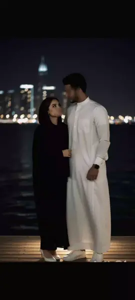 #ك  Create a picture of a young man wearing a white Arab dishdasha, white sneakers, and a gray wristwatch. The girl is wearing a black abaya and a black shawl with light makeup, beautiful eyeliner, and simple white high heels. The height difference between them is beautiful, as the young man appears taller than the girl and the girl has a beautiful body. They are standing on a pier near the sea. Behind the sea are the city lights. It is night and the lighting is beautiful. The young man looks at the girl with love and tenderness, so that her full height appears in the picture, without changing any of her features. 