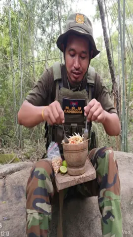 A Cambodian soldier sitting on a rock in a peaceful forest, wearing a camouflage uniform and military vest with a “Cambodia” patch, smiling while eating traditional Khmer papaya salad (bok lahong) from a wooden mortar. He holds a fork in one hand and the mortar in the other. On the small wooden table in front of him are peanuts and lime. The background is full of bamboo trees and natural greenery, warm daylight filtering through the leaves, cinematic and ultra-realistic photography style.