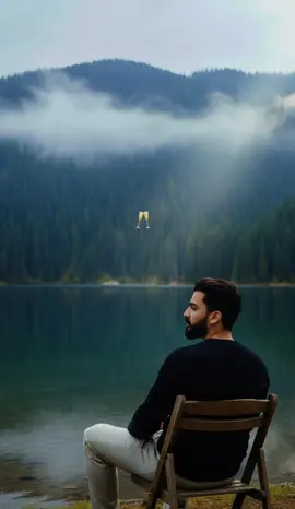 Google Gemini Ai Editing Prompt (Copy Paste)👇🏻 Create Hyper Realistic Photograph Of A thoughtful young man (from uploaded image) with same hair, wearing a  black long-sleeved shirt and light-colored pants, sitting on a camping wooden chair at the edge of a calm lake. He is looking off to the side, A dense, mist-shrouded evergreen forest covers the mountains in the background. Overcast, moody atmosphere, natural light rays coming from top right, cinematic, outdoor adventure. --ar 9:16 --style raw