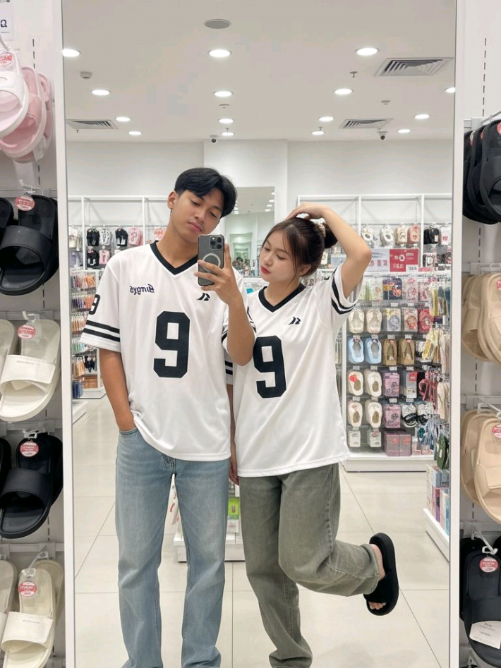 𝙋𝙍𝙊𝙈𝙋𝙏 : Buatkan gambar (saya dan wanita di atas) berswafoto di depan cermin di sebuah toko modern yang terang dengan rak-rak yang tertata rapi dan pencahayaan yang lembut. Saya dan wanita di atas mengenakan pakaian kasual yang stylish.  Wanita di atas mengenakan kaus oversize bergaya sporty berwarna putih dengan detail hitam, terdapat angka sangat besar 