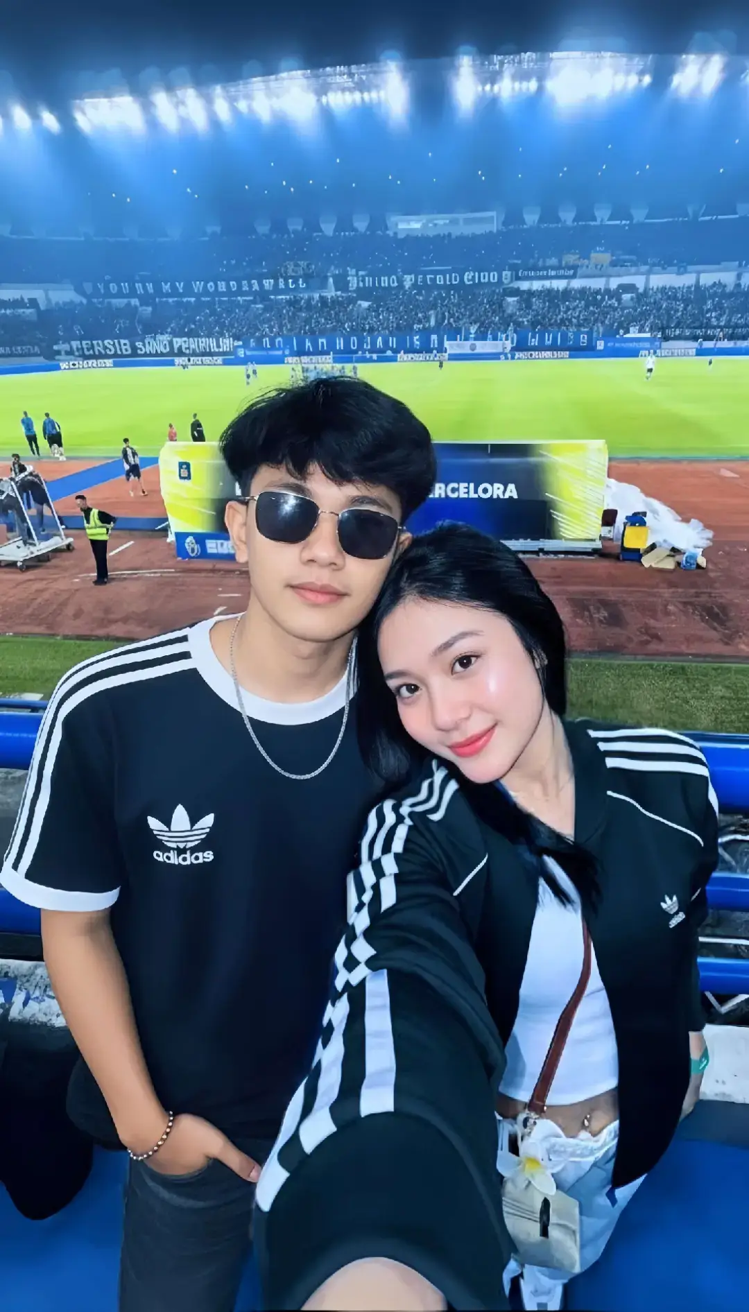 TUTORIAL?? BACA CAPTION‼️ 1. Buka google/apk gemini di hp kalian 2. Masukan photo wajah kalian dan pastekan  prompt:👇🏻 Coba buatkan sebuah foto selfie vertikal dengan komposisi close-up yang romantis dari dua subjek diatas, diambil pada malam hari di tribun sebuah stadion sepak bola yang disinari lampu sorot berwarna biru. Foto ini menangkap sepasang kekasih muda yang berpose serasi dan penuh gaya, dengan kontras pencahayaan yang dramatis. Subjek dan Gaya Berpakaian (Couple Outfit) Pria (Kiri): Memiliki potongan rambut undercut dengan gaya messy atau blow-out yang modern, berwarna hitam pekat. Wajahnya tertutup kacamata hitam dengan bingkai kotak tebal yang menambah kesan edgy dan cool. Ia mengenakan kaus T-shirt hitam berlengan pendek dengan kerah dan ujung lengan yang bergaris putih, serta logo Trefoil Adidas berwarna putih yang tercetak jelas di dada. Ia melengkapi penampilannya dengan kalung rantai perak tipis dan gelang rantai di pergelangan tangan kirinya. Ia berpose dengan tangan kanan dimasukkan ke saku celana panjang gelapnya (mungkin jeans hitam), memancarkan aura santai dan keren. Wanita (Kanan, Pengambil Foto): Berada di sisi kanan frame dan sedikit lebih maju, ia memiliki rambut hitam lurus panjang yang terurai ke depan. Ia mengenakan track jacket hitam dengan three-stripes putih tebal yang khas di bahu dan lengan, di atas crop top tangki putih. Gaya ini sangat serasi dengan pakaian pasangannya. Ia tersenyum lembut dan menatap langsung ke kamera. Stylenya yang modis diperkuat dengan tindik pusar (belly button piercing) dan gantungan kecil berbentuk bunga putih yang tergantung di celana pendek/rok berwarna terang. Posenya sangat intim, menyandarkan kepalanya dengan lembut di bahu pria.  Latar Tengah (Pinggir Lapangan): Di belakang pasangan, terlihat pagar pembatas tribun dan banner LED besar yang terang benderang. Papan reklame ini menampilkan logo sponsor yang mencolok (termasuk tulisan 'PERSIB BANDUNG'' dan slogan lain seperti 'BANDUNG KEMBALI BERGELORA'). Di area track atletik yang berwarna oranye/cokelat, terlihat siluet beberapa staf lapangan (dengan rompi kuning) dan pemain berjersey biru. Latar Belakang Jauh (Tribun Atas): Tribun penonton di seberang lapangan terlihat penuh sesak, dipenuhi oleh kerumunan suporter yang didominasi atribut berwarna biru. Spanduk-spanduk besar yang dibentangkan menciptakan suasana match day yang bersemangat. Cahaya lampu sorot stadion (floodlights) yang sangat kuat mendominasi, memberikan tone warna biru-dingin (cool tone) yang intens pada seluruh latar belakang. Cahaya ini menciptakan efek yang dramatis dan menonjolkan suasana malam. Pencahayaan Subjek: Wajah pasangan diterangi oleh sumber cahaya yang sangat dekat dan lembut (kemungkinan selfie flash atau lampu ponsel), membuat kulit mereka terlihat flawless, cerah, dan glowing (highlighter terlihat jelas di tulang pipi wanita). Pencahayaan ini menghasilkan pemisahan yang jelas dan tajam antara subjek yang terang dan latar belakang stadion yang gelap dan kebiruan, menempatkan fokus 100% pada ekspresi dan couple outfit mereka. Kualitas Gambar: High resolution dengan fokus yang tajam pada subjek, menangkap detail pakaian dan aksesoris mereka. 3. Tunggu hasilnya 4. Kalo sudah sesuai Download nahh sekarang kamu udh ngga bakal dihina lagi Bree👍🏻 #prompt #geminiai #persib #bandung #casualstyle 