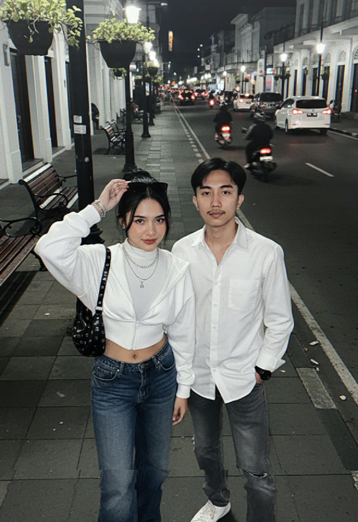 prompt Gemini ai di jl Malioboro bareng pasangan terbaru. Buat gambar potret pria dan wanita wanita (orang di foto yang diunggah), pertahankan wajahnya 10000% sama dengan foto yang diunggah, dengan mata, alis, hidung, dan mulut dipertahankan 1000%. Rambut pria sama seperti yang di foto dan wanita mengenakan Detail Karakter Pakaian Atasan: Cropped Zip-Up Hoodie Jaket Tudung Ritsleting Pendek) tambahkan inner baju berwarna putih agar tidak terlihat leher dan pusar. Warna: Putih. Bentuk/Panjang: Model oversize setinggi pinggang, menonjolkan bagian pinggang dan perut. Lengan: Lengan panjang. Leher/Atas: Memiliki tudung (hoodie) dan ritsleting depan yang diturunkan, membentuk leher V yang dalam. Desain: Ketatkan (body-hugging), menonjolkan lekuk tubuh. Mungkin memiliki detail jahitan Atau garis-garis yang membantu atasan terlihat lebih pas seperti korset di area tubuh. Bawahan: Celana Jeans Jenis: Celana jeans longgar (loose-fit) dengan potongan kaki lebar (flared), mengikuti tren Y2K yang populer. Bentuk: Bagian kaki celana sangat lebar, mulai dari paha hingga ujung kaki. Warna/Warna Luntur: Warna luntur (wash) biru tua/medium dengan efek luntur yang terlihat alami. Aksesori Kalung: Mengenakan kalung berlapis (layered) yang menonjolkan logam perak. Perhiasan: Beberapa gelang dan cincin di jari-jari dan pergelangan tangan. Tas: Tas selempang bahu (shoulder bag) berwarna hitam model Baguette. Paku Keling/Kancing: Logam. Sepatu: Sepatu olahraga/sneakers. Warna: Putih/Perak. Detail Tata Rias, Rambut, Kamera, dan Pencahayaan 59 Tata Rias (Makeup) Riasan: Nuansa alami (natural tone) yang memberikan kesan manis. Wanita memiliki rambut yg di ikat asal dan sedikit berantakan sehingga anak rambut dan akar dasarnya ny menutupi wajahnya yang memberi kesan manis dan imut . Kulit: Putih, mulus, dan cerah. Pipi: Perona pipi (blush on) warna oranye kemeraha Pria mengenakan kameja putih, celana abu² dengan sobekan dan sepatu sneakers putih Lokasi: Trotoar di jalan Malioboro yogyakarta pada malam hari. Struktur Atas ujung: Terdapat struktur  bangunan bergaya kolonial, deretan bangku besi, serta pot tanaman gantung di tiang lampu di atas kepala, memberikan bayangan ke permukaan di bawahnya. Permukaan Jalan: Jalan beraspal berwarna abu-abu gelap. Trotoar: Area tempat pria dan wanita tersebut berdiri  Suasana: Memberikan nuansa gaya perkotaan di malam hari yang agak mentah (raw) dan menonjolkan struktur beton. Di latar belakang terdapat taksi, pengemudi sepeda motor, dan mobil mewah yang diambil dengan efek blur pergerakan, menambahkan dimensi dan nuansa kota yang hidup. Posisi Berdiri fokus hanya pada pria dan wanita. Posenya pria dan wanita  berdiri santai di trotoar Jalan Malioboro pada malam hari, di bawah papan jalan berwarna hijau bertuliskan 