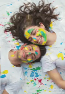 Gemini prompt : 🎨🫟🖌️  Estetika: Ramah, tulus, ceria, artistik, bersemangat, dan sedikit berantakan. Pencahayaan: Cahaya siang yang lembut, alami, dan menyebar, mungkin mendung atau sedikit teduh untuk menghindari bayangan yang tajam dan memungkinkan warna menonjol. Cahaya harus menerangi wajah secara merata dan mendistribusikan cat secara merata. Palet Warna: Warna-warna cerah, primer, dan sekunder (kuning, biru, merah, hijau, putih) mendominasi cat dengan latar belakang kontras warna kulit dan kain putih. Subjek: Dua orang (dewasa muda) berbaring: Mereka saling berhadapan. Kepala mereka berdekatan, menunjukkan kedekatan atau momen kebersamaan. Berlumuran cat: Kedua orang tersebut memiliki cat warna-warni yang cukup banyak di wajah, rambut, dan pakaian mereka (kaos/pakaian putih). Aplikasi cat bersifat spontan dan ceria, tidak presisi. Ekspresi: Orang di atas (perempuan) tertawa terbahak-bahak, matanya terpejam gembira, dan giginya terlihat. Orang di bawah (maskulin) memiliki senyum lembut, penuh kasih sayang, dengan mulut tertutup, mata terbuka lembut, dan menatap orang lain. Rambut: Keduanya memiliki rambut yang kusut karena cat. Rambut orang feminin terurai; rambut orang maskulin lebih pendek. Detail: Tekstur Cat: Cat harus memiliki tekstur yang agak kental, basah, dan bercak-bercak, yang menandakan cat masih baru atau baru diaplikasikan. Pakaian: Kain putih (seperti kaus atau seprai) berfungsi sebagai kanvas untuk cipratan cat yang lebih banyak dan menonjolkan kekacauan warna-warni. Aksesori: Anting-anting kecil dan cantik yang terlihat pada orang feminin. Kalung atau rantai yang halus. Komposisi: Sudut: Bidikan sudut tinggi yang melihat langsung ke objek. Bingkai: Bidikan yang rapat dan intim yang berfokus pada tubuh bagian atas dan wajah dua orang. Komposisi: Objek-objek tersebut berdempetan, tubuh mereka membentuk sedikit lengkungan atau bentuk 