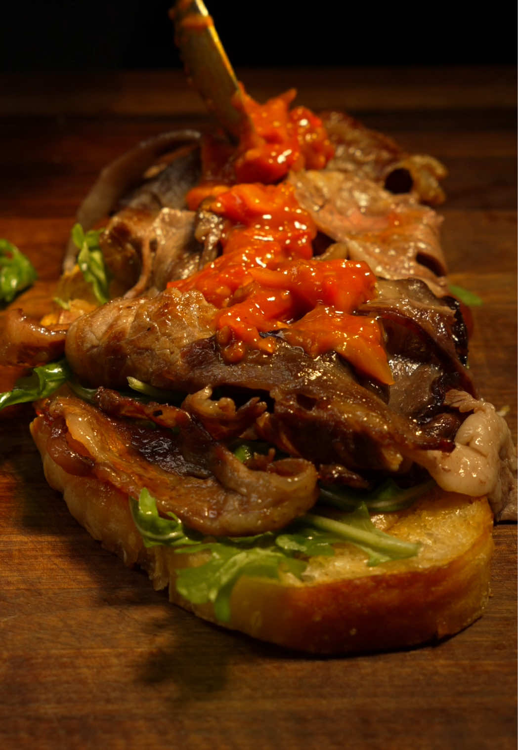 Ribeye Steak Sandwich. When I think of a steak sandwich, I usually imagine a beautiful steak cooked medium rare, sliced up between bread with all the works. But I recently got a new kitchen gadget, a meat slicer, and thought I’d try shaving the steak instead to create thin, delicate layers of meat. To start, I left a prime grade ribeye uncovered in the refrigerator for two days to let some moisture evaporate and firm it up. That made it easier to get thin, even slices. The slicer cut through it like butter, and I was already getting excited about all the things I could do with this machine. From a one inch thick steak, I ended up with ten perfect slices. My wife was baking fresh sourdough, so I decided to use that for the sandwich. She did all the work, I just finished it for the video, so all credit to her for that incredible bread. I made some confit garlic, though it went a little too far for my liking. I used some of the oil to brush the sourdough slices and toasted them until crisp on both sides. I picked out the garlic pieces that hadn’t browned too much and spread them over the toast. Next, I added a layer of arugula, lightly oiled and seasoned. For the steak, I fired up the griddle. These thin slices cooked fast. Some I seared longer for a nice crust, and others I just kissed the surface for a light sear. I made a tomato and pepper relish by sautéing shallots, cherry tomatoes, and a mix of hot and sweet peppers until they cooked down. Then I added a bit of sugar, a few cracks of black pepper, a splash of red wine vinegar, and finally some anchovies to give it some umami.  I piled the steak slices over the greens and spooned the relish generously on top. Then I spread Kewpie mayonnaise on the top slice of bread, pressed it all together, sliced it in half, and served it with a side of curly fries. The sandwich was fantastic. The flavor was very roast beefesque, but better. I love a classic rare steak sandwich, but this style is definitely up there. Still, a few tweaks would take it to a solid 9. A touch of whole grain mustard, a bit more meat (I used six slices but next time I’ll go with all ten), and turning that confit garlic into an aioli instead of spreading it plain just to take that edge off. 