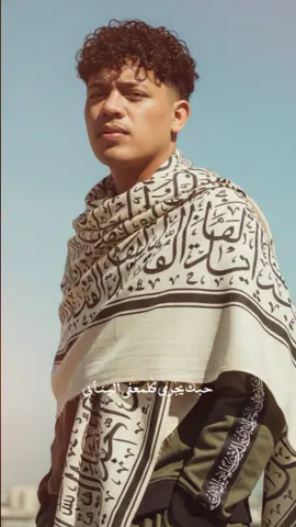 Promt nya Foto menggunakan wajah asli saya, buatkan gambar potret dengan konsep sebagai berikut: Potret editorial bergaya majalah Vogue yang menampilkan seorang pria muda dengan gaya elegan dan klasik. Subjek berdiri dengan ekspresi percaya diri, menatap ke arah kamera dengan sedikit squint karena cahaya matahari. la mengenakan syal besar atau selendang berwarna krem muda/beige dengan pola huruf kaligrafi Arab berwarna hitam yang menutup sebagian bahunya. Di balik syal tersebut, ia mengenakan pakaian berwarna hijau zaitun gelap atau army green, dengan detail strip hitam dengan kaligrafi Arab di sepanjang lengan. Kombinasi pakaian ini menciptakan perpaduan gaya streetwear modern dan elemen budaya. Tangan kanannya dimasukkan ke saku. Latar belakang menampilkan langit biru cerah dengan sedikit kabut lembut dan bentuk samar bangunan kota di kejauhan, memberikan kesan suasana urban pada siang hari yang cerah. Warna dan tone keseluruhan gambar mengarah pada vintage aesthetic, dengan saturasi lembut dan kontras hangat khas majalah era 80-90-an. Pencahayaan: pencahayaan alami dari sinar matahari langsung, membentuk bayangan tegas di sisi wajah dan tubuh subjek, menciptakan efek dramatis dan depth yang kuat. Pengambilan gambar: diambil dari angle slightly low shot (sedikit dari bawah), framing medium shot dari kepala hingga paha, memberikan kesan dominan dan elegan. Fokus tajam pada subjek, dengan latar belakang blur lembut (shallow depth of field). Suasana: gaya editorial berkelas dengan nuansa retro, memancarkan aura maskulin, tenang, dan artistik. Foto ini mencerminkan kombinasi antara fashion klasik dan modern minimalis, cocok untuk cover atau spread majalah high-fashion seperti Vogue. #CapCut #moots? #arabicsong 