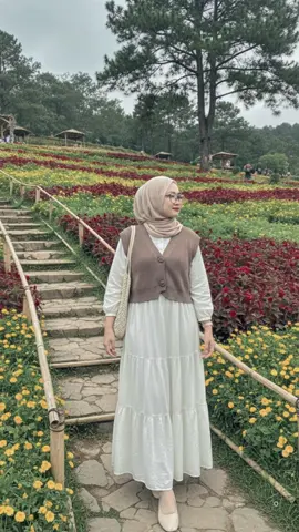 PROMPT GEMINI AI👉🏻. Edit foto ini menjadi seorang wanita muda berhijab yang berdiri santai di sebuah taman bunga yang luas dan terawat, menciptakan suasana yang sejuk dan asri. Pose: Wanita tersebut berdiri dengan tubuh menghadap sekitar tiga perempat ke kanan, dan kepalanya menoleh ke arah kamera dengan pandangan sedikit keatas, seolah sedang menikmati pemandangan di kejauhan. Wajahnya menunjukkan ekspresi santai dan tersenyum tipis. Arah pandangannya tertuju ke bawah. Tangan kanannya sedikit terangkat ke arah hijabnya, mungkin sedang merapikan atau sekadar gesture santai. Tangan kirinya memegang tas jinjing yang ia kenakan di bahu kirinya. Pakaian (OOTD): Wanita tersebut mengenakan pakaian yang modis dan berlapis, cocok untuk suasana di luar ruangan yang sejuk: • Hijab/Jilbab: Mengenakan hijab pasmina polos berwarna krem muda atau nude dengan gaya yang sederhana dan rapi, membingkai wajah dengan lembut. • Atasan: Mengenakan kemeja lengan panjang berwarna putih sebagai lapisan dasar. • Rompi: Dilapisi dengan rompi rajut tanpa lengan berwarna cokelat tanah atau taupe di atas kemeja putih. Rompi ini memiliki detail kancing di bagian depan dan memberikan tekstur serta kontras warna yang menarik. • Bawahan/Rok: Mengenakan rok panjang (maxi skirt) berwarna putih gading yang mengembang (flowy). Rok tersebut memiliki detail berlapis (tiered) dengan sedikit tekstur atau renda pada bagian pinggang atau lapisan atasnya. • Aksesoris: Membawa tas jinjing (tote bag) berwarna krem atau putih gading dengan tekstur seperti rajutan atau anyaman di bahu kirinya. Ia juga memakai kacamata bingkai bulat hitam. Sepatu yang dikenakan adalah flat shoes berwarna krem muda. Latar Belakang (Lanskap): Latar belakang didominasi oleh lanskap taman yang terawat: • Bunga: Terdapat bidang-bidang bunga yang ditanam dalam barisan memanjang menanjak (terasering) di belakang subjek. Warna bunga yang mencolok meliputi merah cerah (maroon/merah tua), kuning, dan hijau dari dedaunan, menciptakan pola warna yang indah. • Pepohonan: Di bagian paling atas dan belakang, terdapat pohon-pohon pinus yang tinggi dan rindang, menunjukkan lokasi ini berada di daerah dataran tinggi atau pegunungan. • Jalur Setapak: Subjek berdiri di atas jalur setapak berbahan semen atau batu yang sedikit menanjak. Di sebelah kirinya terdapat tangga alami yang dibuat dari bilah-bilah bambu atau kayu yang ditata secara informal, menambah kesan alami pada taman. • Suasana: Suasana tampak sejuk dan asri, seperti di kawasan wisata alam atau agrowisata yang terawat. Ada beberapa orang di kejauhan, menambah kesan tempat wisata yang ramai. Pencahayaan dan Warna: Pencahayaan memberikan kesan cerah namun lembut: • Pencahayaan: Gambar tampaknya diambil pada siang hari dengan cahaya alami yang tersebar (diffused light), kemungkinan saat cuaca sedikit berawan atau subjek berada di posisi yang tidak terkena sinar matahari langsung yang terik. Hal ini menghasilkan bayangan yang lembut dan tidak terlalu tajam. • Kecerahan: Gambar memiliki tingkat kecerahan yang baik, membuat warna-warna bunga dan pakaian terlihat hidup dan jernih. • Warna Dominan: Warna-warna dominan adalah putih dan krem pada pakaian, diimbangi dengan warna cokelat pada rompi, serta warna-warna cerah merah dan kuning dari hamparan bunga yang mencolok. Warna hijau dari dedaunan dan pohon pinus memberikan keseimbangan visual yang menenangkan. Jangan ubah wajah sedikitpun.  #aiprompt #geminiai #promptgeminiai #prompt #fyp 