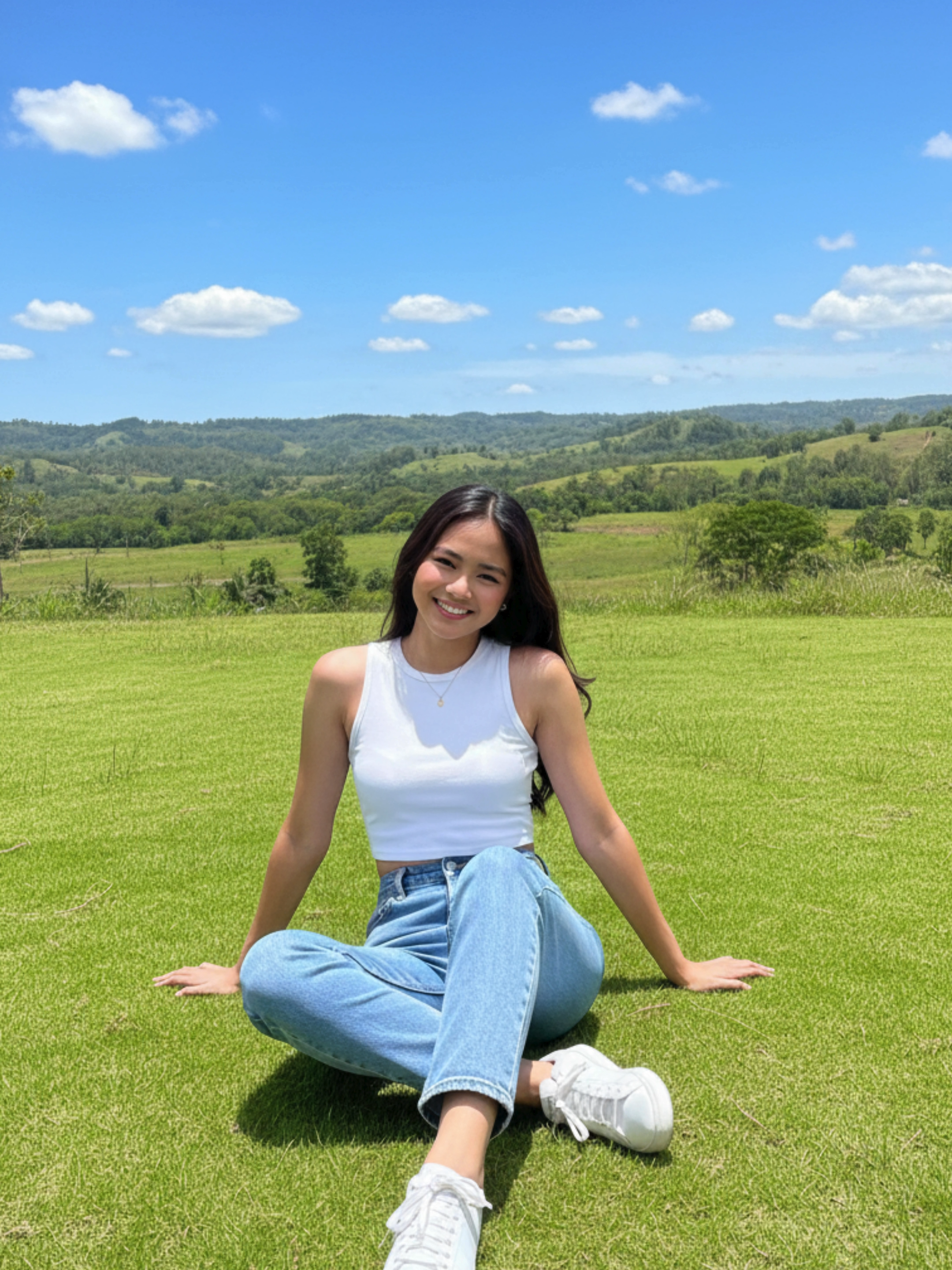 Gemini AI prompt: Three-quarter length outdoor portrait of me (preserve my facial features i should be recognizable) sitting on a grassy lawn in bright midday sunlight, photographed on iPhone 15 Pro using the main wide camera, with no background blur. She wears a white sleeveless crop top and high-waisted blue Levi’s jeans, with white sneakers. she sits at ease with her left arm extended behind and her right hand resting on her crossed right knee. The background features rolling green hills, light patches of woodland, and a vibrant blue sky with scattered small clouds, capturing a serene rural landscape in the Philippines such as around Tagaytay or Rizal. The image displays crisp natural color, clear detail, and high dynamic range typical of iPhone computational photography, rendering both subject and environment sharply and authentically, with all elements in focus and no artificial depth effect.Do not over-edit or enhance the face or expression in any way. Maintain a high degree of facial fidelity to the provided image. avoid background blur
