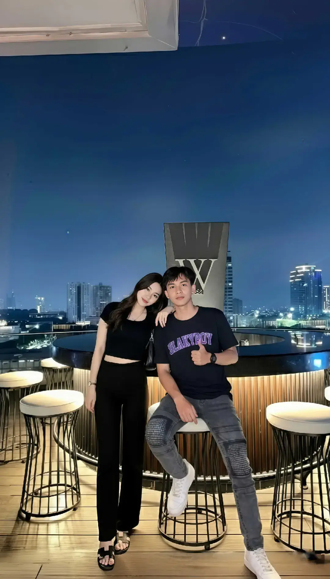 TUTORIAL BACACAPTION‼️ 1. Buka google/apk gemini di hp kalian 2. Masukan photo wajah kalian dan pastekan  prompt:👇🏻 Coba buatkan sebuah foto malam berkualitas tinggi dengan komposisi yang seimbang dan detail visual yang kaya, menangkap momen elegan dari sepasang pasangan muda yang berpose di teras atap (rooftop) sebuah gedung tinggi dari dua subjek diatas. Gambar ini menampilkan dua individu sebagai subjek utama: seorang perempuan muda yang modis dan seorang laki-laki muda yang trendy. Perempuan di sisi kiri mengenakan crop top hitam lengan pendek yang ramping dan celana panjang hitam, menonjolkan siluetnya. Rambut panjangnya yang gelap jatuh bergelombang dengan lembut di pundak, dan ia berpose dengan anggun, tangan kirinya bertumpu di pinggul, memancarkan kepercayaan diri yang tenang. Di sampingnya, laki-laki muda di sisi kanan mengenakan kaus hitam kasual bergambar tulisan 'SKYBOY' yang kontras, dipadukan dengan celana jins abu-abu gelap yang memiliki detail robekan dan patch bergaya. Ia terlihat santai namun bergaya, dengan tangan kanan terangkat sedikit, jempolnya mengacung ke atas, menyiratkan persetujuan atau vibe yang positif. Keduanya berdiri tegak, menghadap langsung ke kamera dengan pandangan yang tenang dan percaya diri, menjadi titik fokus utama yang sangat tajam dan jelas di tengah bingkai. Latar belakang menampilkan panorama cityscape yang memesona, dengan deretan gedung-gedung pencakar langit yang menjulang tinggi, bermandikan ribuan lampu kota yang berkelip-kelip, menciptakan efek bokeh yang lembut di kejauhan, menambah kedalaman dan kemegahan. Langit malam di atasnya adalah hamparan biru tua yang dalam dan pekat, menggambarkan suasana larut malam yang damai namun berenergi. Di antara subjek, sedikit di belakang, berdiri sebuah struktur vertikal gelap dengan bentuk geometris yang unik, menyerupai huruf 'W' besar atau sebuah monumen modern. Latar tengah foto ini didominasi oleh desain interior teras atap itu sendiri: sebuah bar melingkar yang futuristik dan bergaya, dilapisi dengan panel-panel kayu vertikal bergaris-garis. Panel-panel ini diterangi dari bawah oleh lampu aksen kuning keemasan yang hangat, menciptakan efek cahaya yang dramatis dan menonjolkan tekstur kayu yang kaya. Di latar depan, tiga kursi bar bertubuh tinggi dengan jok bundar berwarna putih bersih dan kaki logam minimalis terlihat secara parsial, menambah elemen komposisi dan menunjukkan lingkungan yang mewah. Lantai teras terbuat dari papan kayu yang rapi, menambah sentuhan alami di tengah arsitektur modern. Pencahayaan dalam foto ini adalah masterful, menangkap esensi suasana malam. Subjek utama diterangi dengan cahaya terang dan langsung dari depan, mungkin dari softbox atau lampu studio tersembunyi, membuat detail wajah, pakaian, dan ekspresi mereka terlihat sangat jelas dan menonjol. Kontras antara subjek yang terang benderang dan latar belakang yang lebih gelap namun diterangi oleh lampu kota menciptakan look yang dramatis dan sinematik. Cahaya kuning hangat dari bar di latar tengah memberikan dimensi tambahan, memisahkan subjek dari latar belakang yang lebih jauh. Keseluruhan gambar memancarkan aura kemewahan modern, kepercayaan diri anak muda, dan kehidupan malam urban yang vibrant dan fashionable. 3. Tunggu hasilnya 4. Kalo sudah sesuai Download nahh sekarang kamu udh ngga bakal dihina lagi Bree @zee asadel  #prompt #geminiai  #fyp #jakarta #zee 
