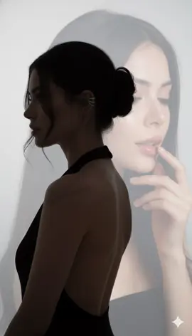 Create a double exposure portrait of a woman in a striking, artistic composition that merges two distinct yet harmonious images into one ethereal vision. In the foreground, there is the silhouette of a woman with jet-black, wavy hair pulled back into a messy bun — her face turned slightly away from the camera, lips parted in a soft, contemplative expression. She wears a minimalist black halter top with a deep, open back, revealing her bare shoulders and elongated neck. A delicate silver ear cuff adorns her left ear, catching the light with a subtle gleam. Her posture is balanced and elegant — arms relaxed, body gracefully angled, as if caught between motion and calm stillness. The lighting is soft and directional, casting gentle shadows that accentuate the contours of her jawline, collarbone, and spine. Overlaid on this figure is a larger, translucent close-up of her face — a dreamlike projection dominating the right side of the composition. This second image shows her with eyes closed, long, defined eyelashes, and slightly parted lips that glisten with a natural sheen. Her skin appears flawless, dewy, and radiant, illuminated by soft, diffused light that enhances her luminous texture. One hand gently touches her lower lip, adding a sense of intimacy and vulnerability. The background is a soft gradient from white to gray, appearing almost empty, allowing the layered figures to stand out with clarity and emotional depth. The contrast between the sharp silhouette and the blurred, glowing face creates a surreal, introspective atmosphere — blurring the line between reality and the inner self. don't change her face #gemini #fyp #f #prompt #ai 