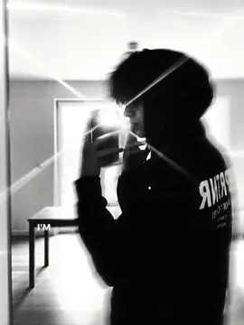 Prompt:create a photo A young man Analyze without changing the reference Tacen taking a selfie in front of the mirror using a mobile phone. He is wearing a black hoodie with a hood that covers part of his hair, posing from the side while holding the phone with one hand. The reflection of the flash light from the phone camera creates a diagonal radiating effect, adding a dramatic impression to the photo. Her hair was dark black and a bit messy, falling on her forehead and covering part of her face. On her hand was one ring on her index finger that gave off an edgy and stylish vibe. Large-sized white writing was seen on the back of the hoodie, creating a strong contrast with the deep black color of the fabric. The background shows the interior of a modern room with white walls, a wooden table, and large windows that emit soft light from the outside. pose: The man stood with his back to the mirror, but his body tilted slightly to the left side. His face is only visible from the side (left profile) as he holds the phone in front of his face for a selfie. The left hand holds the phone in front of the face in a rather high position (at eye level). The fingers form a relaxed yet strong position - partially covering the lower part of the face. The right hand is not clearly visible, it is likely to be on the side of the body. Her shoulders rose slightly because of the position of the hand lifting the phone. The overall pose looks relaxed but stylish, bringing out a cool, mysterious, and slightly edgy aura. Lighting style: black and white (monochrome), high contrast, with light flare effect from the camera. Photo shades: mysterious, cool, minimalist, modern, and urban streetwear style. #geminiai #ai #fyp #trendai #trend 
