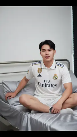 : A highly detailed and realistic digital photograph of a young East Asian man with smooth(Use the face from the reference image.), fair skin sitting cross-legged on a bed inside a modern, minimal bedroom. He has a lean body, athletic build and a clean, symmetrical face with soft, androgynous features. His hair is comma two block korean style, he is wearing a white Real Madrid football kit — jersey and shorts — with gold and navy blue trim, featuring clearly visible team logos, sponsor text, and FIFA Club World Cup badge on the chest. The shorts are slightly loose-fitting. He also wears light-colored ankle socks. His legs are positioned in a relaxed cross-legged pose, with visible light body hair on his thighs.He sits casually on a bed with metal railings and silver-gray satin sheets and pillows. His left arm is extended back, bracing against the bed, and his right hand holds a small object (a car key fob or small device) while resting on his knee. He maintains a relaxed, playful expression with lips slightly parted, his tongue slightly sticking out, looking directly at the camera with a calm gaze.The room features plain white walls, minimal decor, and a black vertical blind partially visible on the side. Lighting is soft, natural or ambient, as if taken in the early evening using an iPhone 13 or 14 front-facing camera in a well-lit room. The overall image is high-resolution with soft shadows and neutral tones, creating a clean and intimate atmosphere.#CapCut #gemini #beach #pantai #pantaivibes 