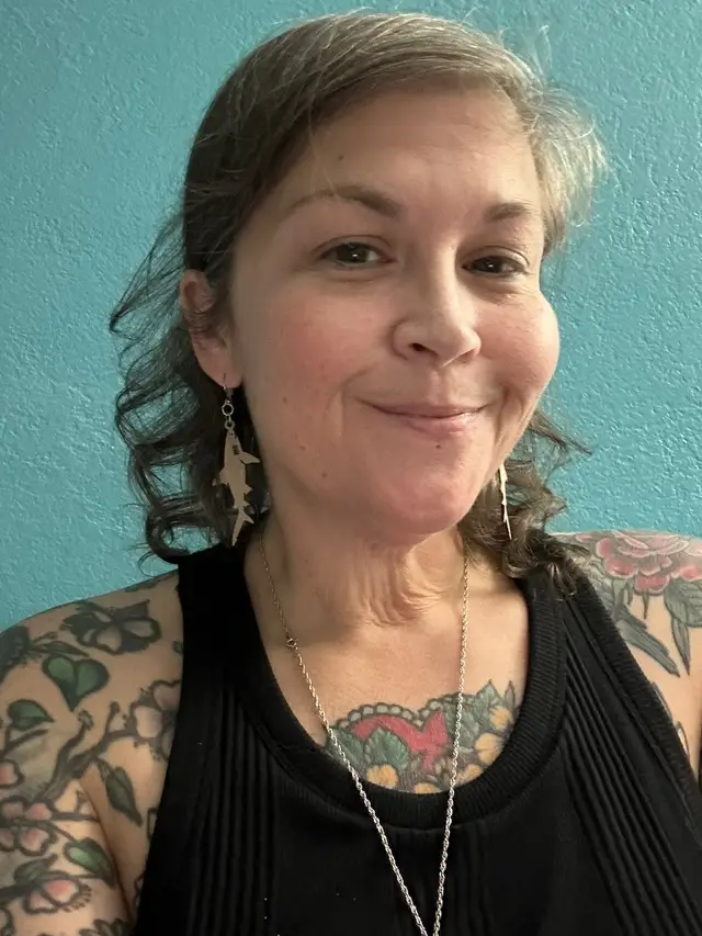 I’ve been feeling beyond stir crazy in Seattle lately, like my skin was so itchy I wanted to unzip and step out of it. So yesterday I made an impromptu decision to take a side quest and this morning I woke up in a place I’ve never been before, on the way to another place I haven’t been before, but is one where some of my ancestors are buried. One of the two places where I have relatives buried in the PNW. I have no idea what to expect, but I’m excited about this journey! Image description: Selfie of Frankie, an olive skinned tattooed human with medium length brown curly hair streaked with silver, smiling and wearing a black tank top and shark earrings. 