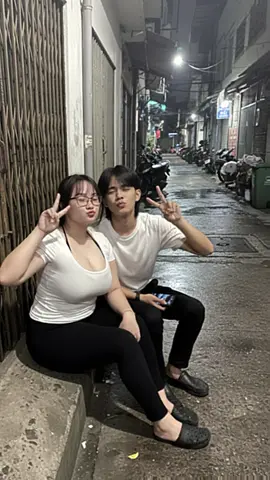 PROMPT GEMINI AI👇 Latar gang malam hari; trotoar basah yang memantulkan cahaya; jalan sempit dengan kendaraan parkir dan tanaman pot yang berserakan; sepasang kekasih pria dan wanita muda (seperti di foto referensi yang saya kirim,wajah tidak di ubah sama sekali) mereka duduk berdeketan di beton rendah di samping gerbang lipat logam. pria: berpose santai dan tersenyum tipis wanita: berpose dengan tanda perdamaian dan ekspresi ciuman palsu; pencahayaan redup, hanya diterangi oleh lampu kilat kamera langsung yang  menyoroti wajah mereka. Latar belakang; pakaian kasual yang terdiri dari atasan putih pas badan; pose duduk santai dengan kaki terentang; ponsel pintar diletakkan di paha; bayangan halus di tanah; atmosfer lembap tersirat oleh kilau pada permukaan; tekstur gerbang logam berkarat dan trotoar yang tidak rata ditekankan. Sudut kamera: 