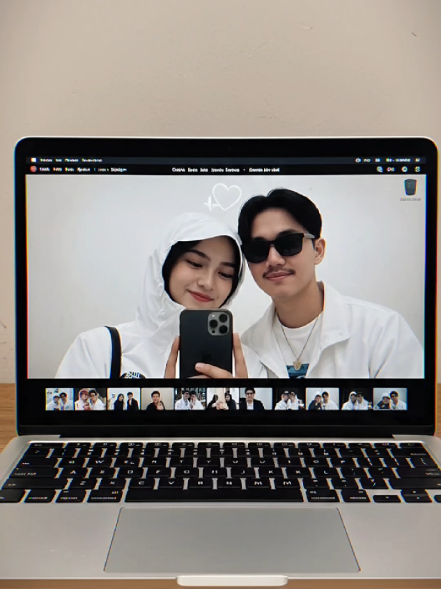 prompt : Subjek 1: Gadis dalam gambar Subjek 2: Pria dalam gambar Momen di belakang layar yang nyaman menunjukkan Subjek 1 dan Subjek 2 mengisi layar laptop saat mereka bersandar berdekatan untuk selfie webcam yang menyenangkan - Subjek 1 di sebelah kiri tersenyum dengan ekspresi playfull dan imut dengan debaran stiker hati putih virtual di atas kepalanya sementara Subjek 2 ekspresi sedikit senyum; keyboard dan bezel MacBook Air membingkai adegan di latar depan, pratinjau thumbnail kecil dari bidikan lain yang melapisi bagian bawah layar dan dinding bertekstur lembut yang terlihat di atas layar laptop membuat komposisi berlapis, sedikit voyeuristik. Subjek 1 memegang ponsel iphone yang digunakan untuk mengambil gambar, jari-jari terlihat pada casing hitam matte ponsel; Subjek 2 memakai pakaian yang sesuai dengan foto terlampir dan kacamata hitam, kedua Subyek berdekatan satu sama lain sehingga tampilan terasa intim dan jujur. Refleksi dan silau pada kaca laptop, cahaya samar kafe interior atau lampu studio di belakangnya, kontrol pengambilan di layar MacBook, dan permukaan desktop yang hangat dan hidup semuanya menambahkan detail sentuhan; bingkai keseluruhan sedikit di luar pusat dengan subjek 
