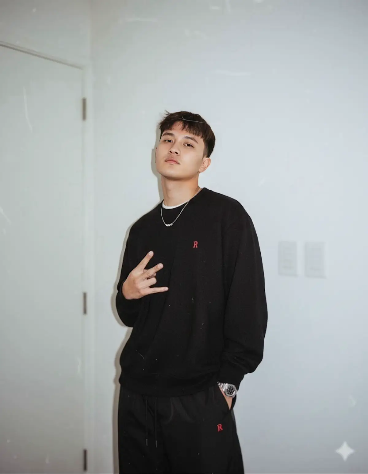 ubah gambar ini tanpa merubah wajah dan rambutnya menjadi potret vintage seolah diambil menggunakan kamera analog saat malam hari,menampilkan seorang pria muda berdiri didalam ruangan yang estetik didekat pintu putih dengan dinding putih polos sebagai latar belakang. ia mengenakan sweater oversized berwarna hitam berlogo R kecil ia menggunakan kalung kecil elegan dengan tekstur lembut dan tangan kirinya dimasukkan ke kantong celana, ia memakai jam tangan silver, ia memakai celana kolor hitam berlogo R kecil warna merah. tangan kanannya membentuk gesture tiga jari seperti pose santai. wajahnya menampilkan ekspresi tenang dan santai. terlihat dua saklar putih disampingnya. pencahayaan: flash langsung yang keras. gaya: vintage gritty ala anak muda tahun 2000- an, warna redup, grain, dan ketidaksempurnaan visual. #gemini #geminitrend #foryoupage #fyp #chatgpt 