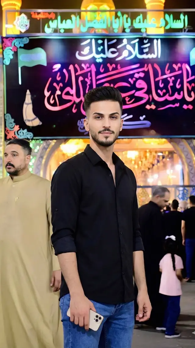 The picture shows a young man standing in front of the staff, wearing a black shirt with raised sleeves and blue jeans, holding a light-colored smartphone in his right hand. The young man looks confident and calm, looking directly at the camera. Background: It is the same as the attached picture. • The background is brightly lit and shows what appears to be the entrance to a religious square or parade, perhaps during an Ashura occasion, where many people wear black clothes. • The upper part of the background is dominated by a large sign with luminous Arabic calligraphy, with religious phrases such as 